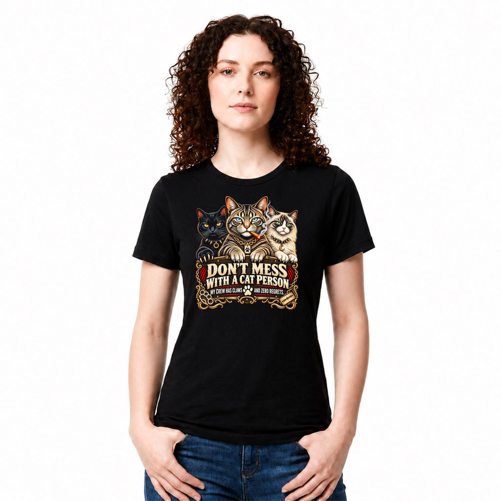 A woman with curly brown hair wearing a black graphic t-shirt with three cats and the text 'DON'T MESS WITH A CAT PERSON' in a vintage western-style design. She is also wearing blue jeans, set against a plain white background.