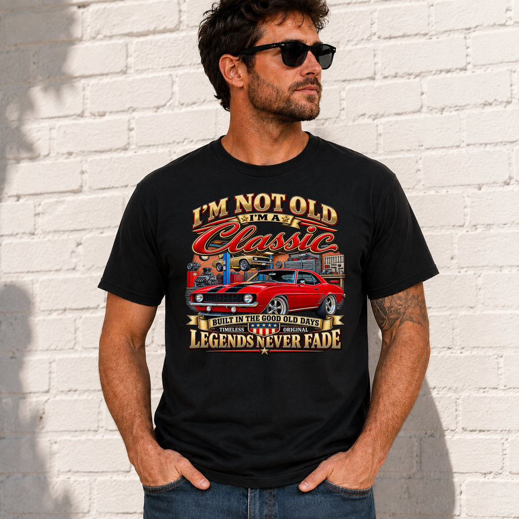 A man wearing a black graphic t-shirt with a red muscle car design and text, standing against a white brick wall.