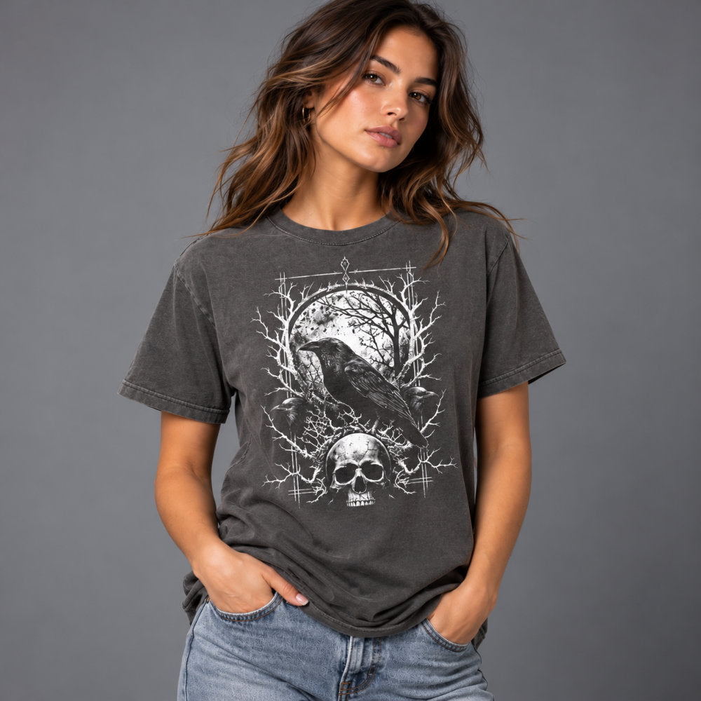 A woman wearing a dark gray ( pepper ) washed graphic t-shirt with a white print of a raven, skull, and tree branches, paired with blue jeans, set against a solid gray studio background.