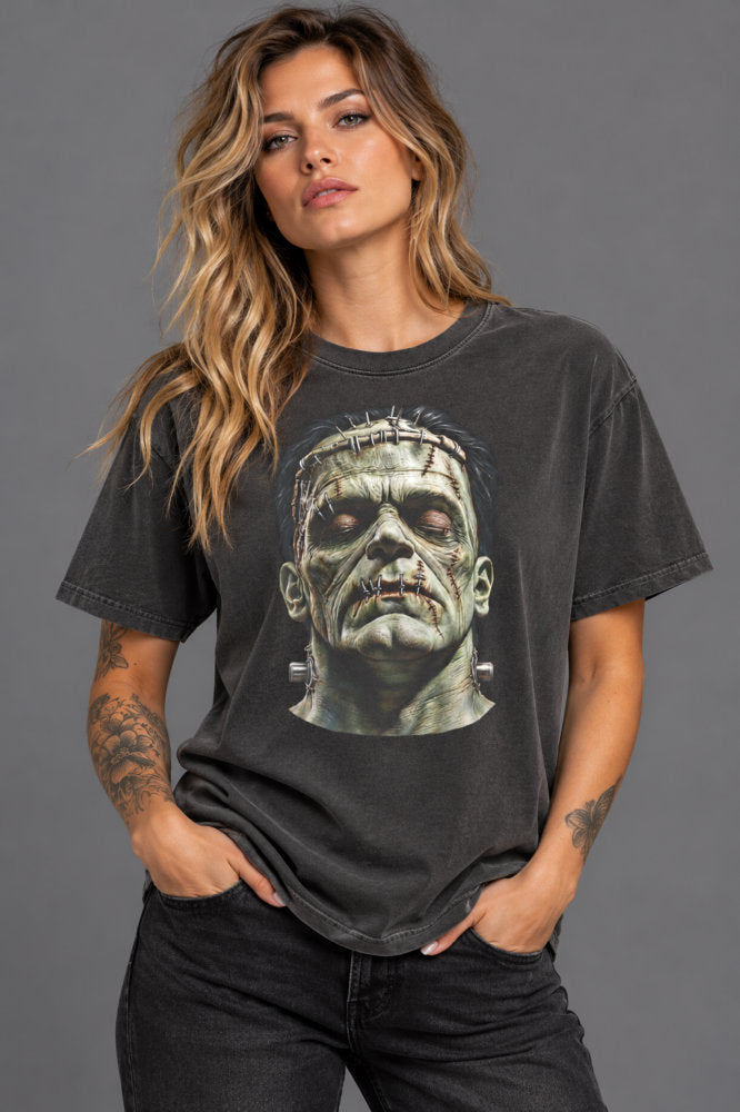 A woman modeling a dark gray washed graphic t-shirt featuring a detailed print of the character Frankenstein's monster, paired with black jeans against a solid gray studio background.