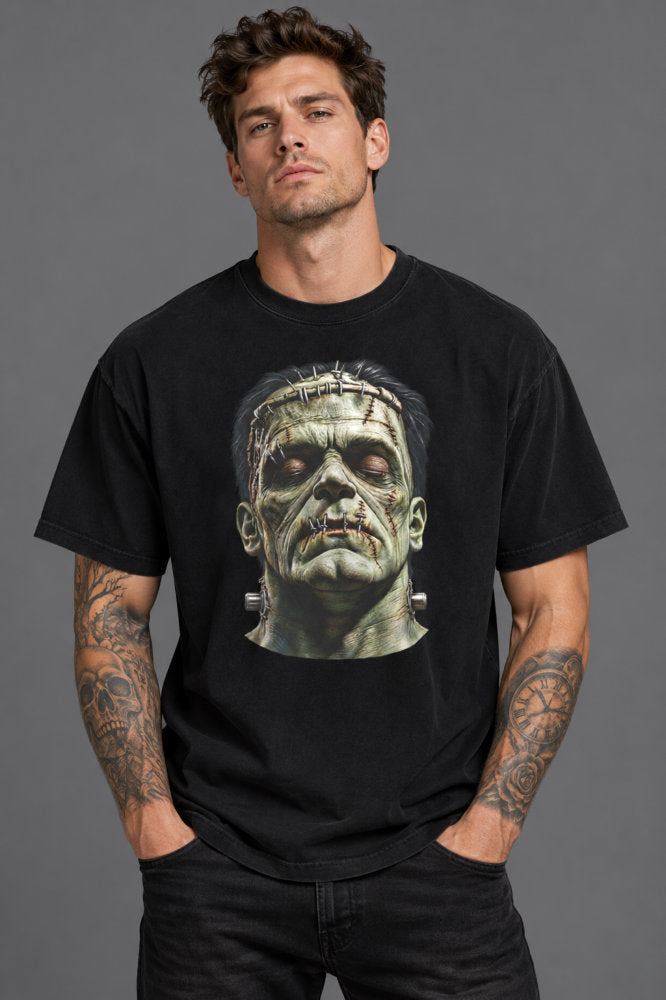 A man wearing a black graphic t-shirt featuring a detailed print of the character Frankenstein's monster head on the chest, posed against a solid gray studio background.