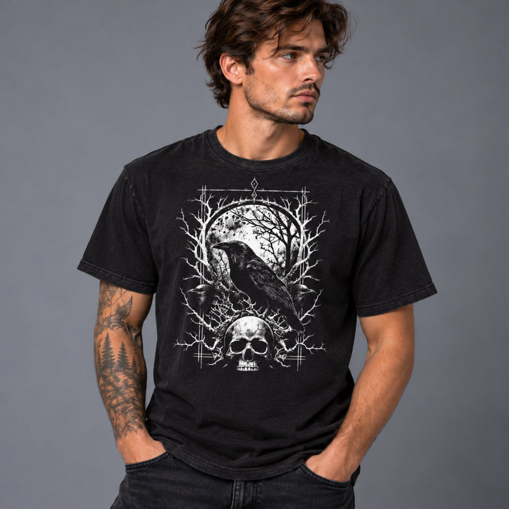 A man wearing a black graphic t-shirt with a white raven, skull, and tree design, posed against a solid gray studio background.