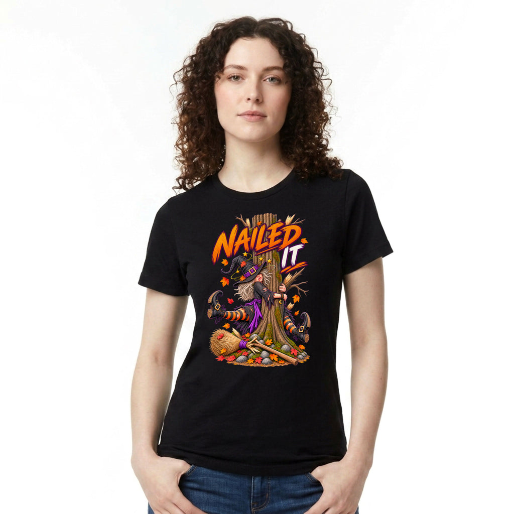A woman with curly brown hair wearing a black short-sleeve graphic t-shirt with a 'Nailed It' witch tree illustration, paired with blue jeans, against a plain white background.