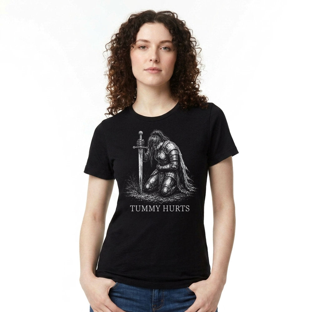 A woman with curly brown hair wearing a black short-sleeve graphic t-shirt and blue jeans, standing against a plain white background. The t-shirt features a white graphic of a kneeling armored figure holding a sword, with the text 'TUMMY HURTS' printed below.