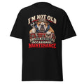 A black short-sleeve crewneck t-shirt featuring a graphic of an bulldog wearing a mechanic's hat and holding a wrench and toolbox, with the text 'I'M NOT OLD I JUST REQUIRE OCCASIONAL MAINTENANCE' in red, white, and beige.